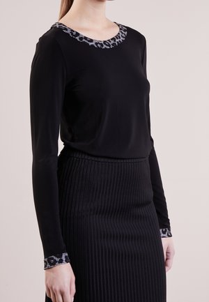 Mujer con un top negro de manga larga con detalles estampados en el escote y los puños, combinado con una falda negra de cintura alta y tejido acanalado.