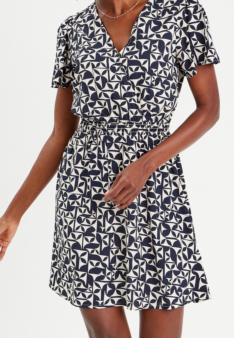 Femme portant une robe à manches courtes, imprimé géométrique noir et blanc, taille élastique et décolleté en V.