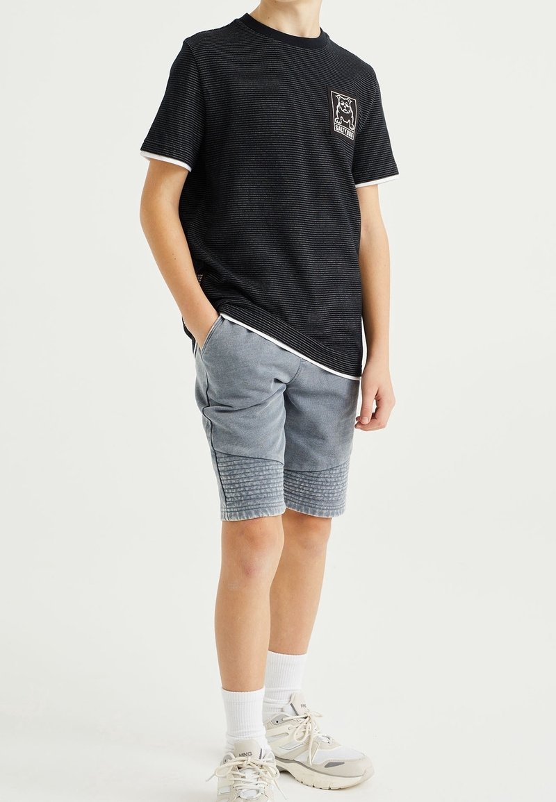 Garçon portant un T-shirt noir rayé avec un logo de chien, un short gris texturé, des chaussettes blanches et des baskets blanches, debout avec une main dans la poche.