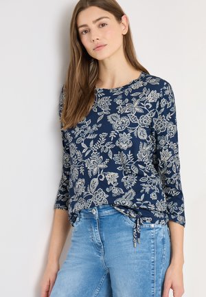 Jeune femme portant un haut bleu marine à manches longues à motifs floraux avec un nœud sur le côté, associé à un jean denim bleu clair, debout contre un mur blanc.