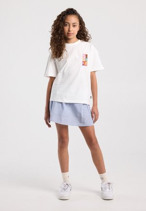 Jonge persoon met krullend haar, gekleed in een wit T-shirt met een kleurrijke patch, een blauw gestreepte rok, witte sokken en witte sneakers, staande tegen een witte achtergrond.
