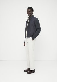 Ναυτικό μπουφάν bomber με κοντράστ γκρι μανίκια, ριμπ μανσέτες, κλείσιμο με κουμπιά τύπου snap, κρεμ πουκάμισο, ανοιχτό γκρι παντελόνι και σκούρα παπούτσια.