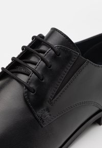 Chaussure en cuir noir à lacets avec une finition lisse, des détails de surpiqûres subtils et un talon bas. Le design présente une forme élégante et minimaliste.