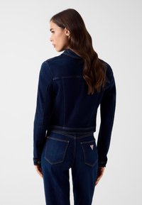 Kurtka dżinsowa w ciemnym niebieskim kolorze, dopasowany krój z klasycznym kołnierzem, widocznym przeszyciem oraz naszywką z logo na tylnej kieszeni. Gładka faktura.