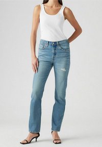 Jean en denim bleu clair à taille haute, design à jambe droite, légèrement usé, et style classique à cinq poches. Associé à un débardeur blanc.