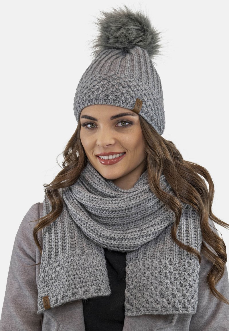 Bonnet en tricot gris avec un pompon en fourrure et une écharpe assortie. Motifs texturés avec une finition tissée. Comprend un petit accent en cuir.