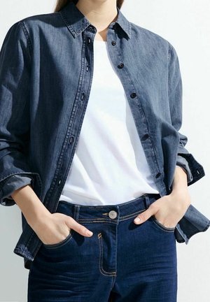 Person iført en mørkeblå denimskjorte over en hvid t-shirt med hænderne i lommerne på matchende mørkeblå jeans.