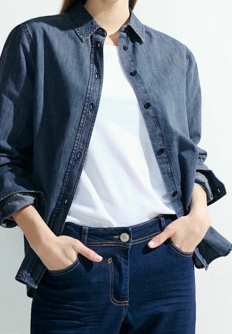 Personne portant une chemise en denim bleu foncé sur un t-shirt blanc, les mains dans les poches d'un jean bleu foncé assorti.