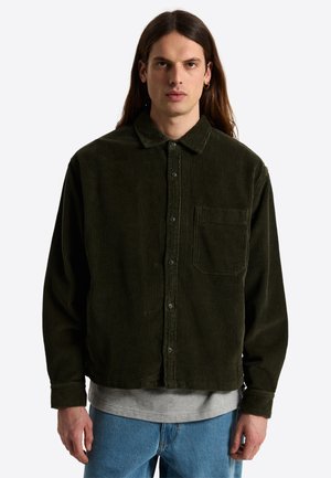 Man met lang bruin haar, die een donkergroen ribfluwelen overhemd met knopen draagt over een grijs T-shirt en blauwe spijkerbroek, staand tegen een witte achtergrond.