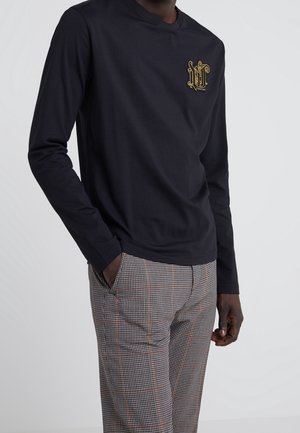 Man draagt een zwart shirt met lange mouwen met een goud geborduurd embleem op de borst en grijze geruite broek met oranje strepen, handen in de zakken.