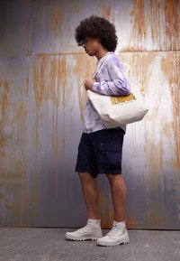 Młody mężczyzna o kręconych włosach, ubrany w koszulę z tie-dye z długim rękawem, granatowe szorty, białe buty, niesie beżową torbę na ramię, stojąc przed zardzewiałą metalową ścianą.