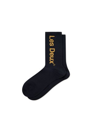 Chaussettes noires côtelées avec le logo "Les Deux" en texte doré sur le côté. Texture lisse, longueur haute à la cheville et design simple.