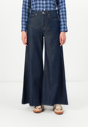 Jean en denim à jambe large de couleur bleu foncé avec une taille haute, comprenant des poches avant et une fermeture à bouton. Associé à des baskets beiges.