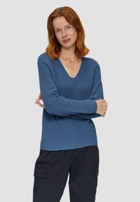 Blauer Strickpullover mit V-Ausschnitt, gerippter Textur und langen Ärmeln. Auffällige seitliche Details umfassen kleine vertikale Perforationen.