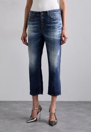 Femme portant un jean court bleu délavé avec fermeture à boutons et des chaussures à talons hauts argentées, debout sur un sol gris.