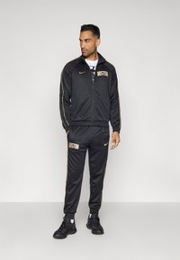 Čierny tréningový komplet Nike so zlatými akcentmi, ktorý obsahuje nápis "BOSTON" na bunde a nohaviciach. Bunda má zips a stojací golier.