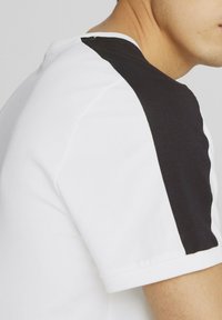 Camiseta de manga corta con un cuerpo blanco y un panel negro en el hombro. Fabricada en suave algodón, con un cuello redondo y dobladillo cosido.
