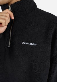 Persona sosteniendo la cremallera de una chaqueta de forro polar negro texturizado con el logo blanco "PEGADOR" en el pecho, frente a un fondo blanco.