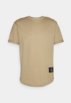 Beżowy bawełniany t-shirt z krótkimi rękawami, okrągłym dekoltem i lekko zaokrąglonym dołem. Na boku znajduje się czarna naszywka z logo CK.