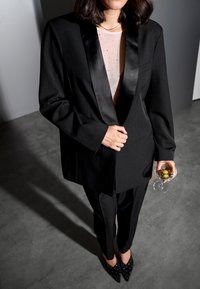 Čierny na mieru šitý oblek s hodvábnym klopovým reverom, priliehavé nohavice, priesvitný bodkovaný top, držiaci pohár s olivami, nosené s čiernymi lodičkami s ihličkovým podpätkom.