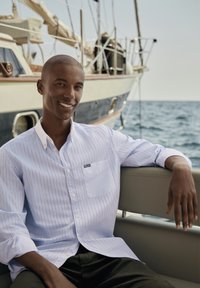 Camicia button-up a righe blu chiaro con colletto e tasca, indossata con pantaloni scuri, su una barca con uno sfondo acquatico.