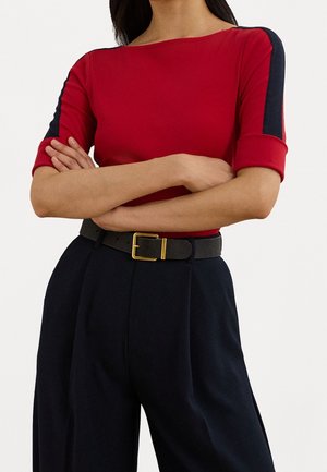 Kobieta ubrana w czerwony top z czarnymi paskami na rękawach, czarne spodnie z wysokim stanem i zakładkami oraz czarny pasek ze złotą sprzączką, skrzyżowane ramiona.