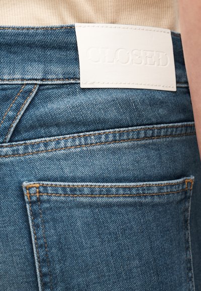 Jeans di denim con una toppa in pelle bianca etichettata "CLOSED" in vita, con tessuto blu e cuciture arancioni visibili.