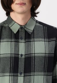 Camisa de cuadros con un patrón verde y negro, hecha de una tela texturizada, con cierre de botones y cuello. Botones negros visibles.