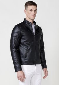 Giacca di pelle nera con spalline trapuntate, tasche con zip e colletto rialzato. Indossata sopra una camicia bianca e pantaloni bianchi aderenti.