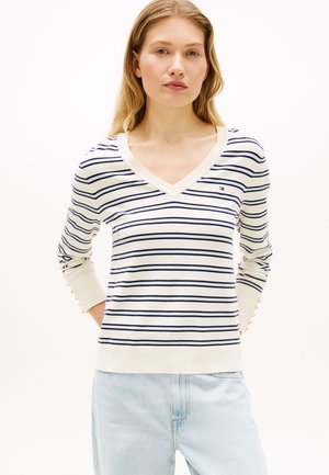 Mujer con cabello rubio largo, vistiendo un suéter blanco de manga larga con cuello en V y rayas horizontales azul marino, y jeans azul claro, de pie contra un fondo blanco.