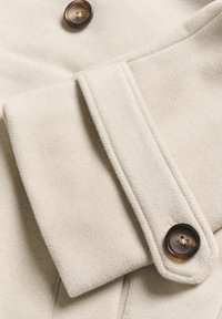 Țesătură de lână de culoare crem, cu o suprafață texturată, având un manșon pliat și un nasture maro închis, cu patru găuri. Elementele de design sunt minimalist.