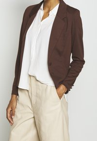 Brun figurerad blazer med kavajkrage, kombinerad med en vit skjorta med knappar och beige vida byxor, som framhäver en mjuk tygtextur.