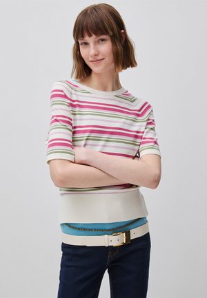 Junge Frau mit kurzen braunen Haaren, die ein gestreiftes Kurzarmtop in Pink, Grün und Weiß, blaue Jeans und einen weißen Gürtel trägt, mit verschränkten Armen.