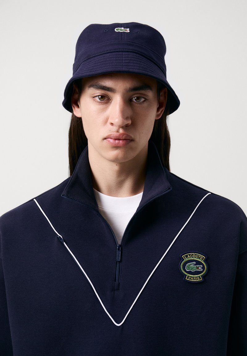 Pull léger navy à demi-fermeture éclair avec un passepoil blanc, présentant un écusson du logo Lacoste. Associé à un chapeau de seau navy assorti, texture lisse.