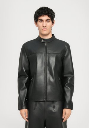 Young man with short curly hair wearing a black leather zip-up jacket and matching black leather pants against a plain background.