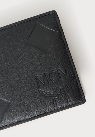 Coin de portefeuille en cuir noir avec logo MCM embossé et emblème ailé, présentant des motifs texturés en forme de losange sur la surface.