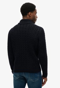 Pull en maille bleu marine avec un col montant et un motif torsadé texturé, poignets et ourlet côtelés, porté avec un jean en denim bleu.