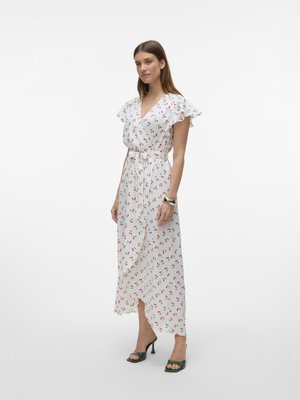 Frau trägt ein weißes Wickelkleid mit rotem Kirschmuster, flatternden Ärmeln und Bindegürtel, kombiniert mit dunkelgrünen Sandalen mit Absatz.