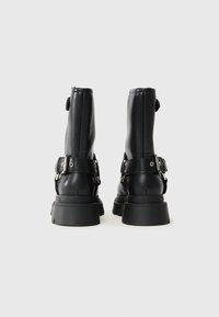 Botas de tobillo de cuero negro con suela gruesa, hebillas laterales y detalles de costura. Presenta una lengüeta en la parte posterior para facilitar su colocación.