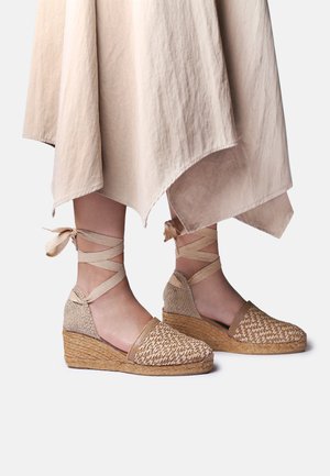 Beige gevlochten sleehak espadrilles met striklinten bij de enkel, gedragen met een lichtbeige asymmetrische zoom rok op een effen achtergrond.