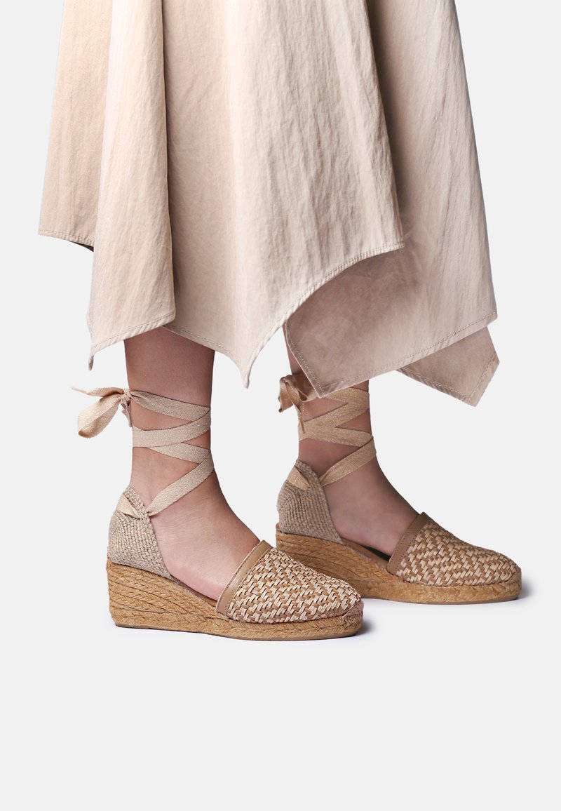 Beige gewebte Keil-Espadrilles mit Schnürbändern am Knöchel, getragen zu einem hellbeigen asymmetrischen Rock vor einfachem Hintergrund.