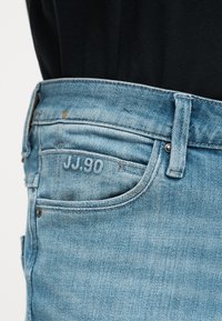 Κοντινό πλάνο σε τζιν παντελόνι από ανοιχτό μπλε denim με μπροστινή τσέπη που δείχνει το κεντημένο λεπτομέρεια "JJ.90" και θηλιά για ζώνη, φορεμένο με μαύρο τοπ.