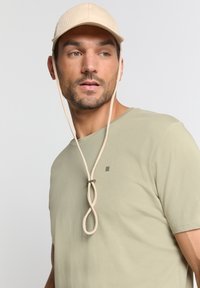 Beige pet met verticale strepen, verstelbare kinband, gecombineerd met een lichtgroene T-shirt met een klein logo. Eenvoudig, functioneel ontwerp.