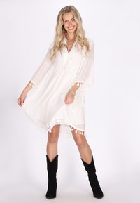 Weißes Kleid mit ausgestellten Ärmeln, fransigem Saum und V-Ausschnitt. Das Model trägt schwarze Wildlederstiefel. Glatte Textur, leichtes Material.