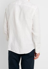 Camicia bianca in lino con una texture liscia, maniche lunghe, polsini con bottone e orlo curvo, abbinata a pantaloni scuri.