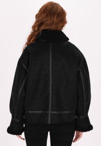 Musta takki, jossa on teksturoitu pinta, lampaanvillakaulus ja kontrastiväriset mustat yksityiskohdat. Ominaisuuksiin kuuluu rento istuvuus ja leveät hihat.