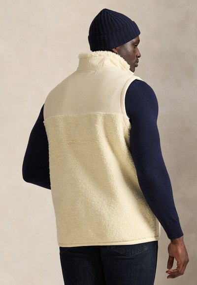 Krämfärgad fleecerväst med ett slätt tyg på överdelen, hög krage och sidosfickor, bärs över en marinblå långärmad skjorta. Mörka jeans fulländar looken.