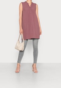 Tunika senza maniche in tessuto mauve con colletto e dettaglio dei bottoni, abbinata a jeans skinny grigi e una borsa beige. Le scarpe hanno un tacco a blocco.