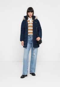 Giacca puffer navy con zip e cappuccio in pelliccia sintetica, maglione a righe marrone e crema sotto, jeans a gamba larga azzurri, e una piccola borsa nera.