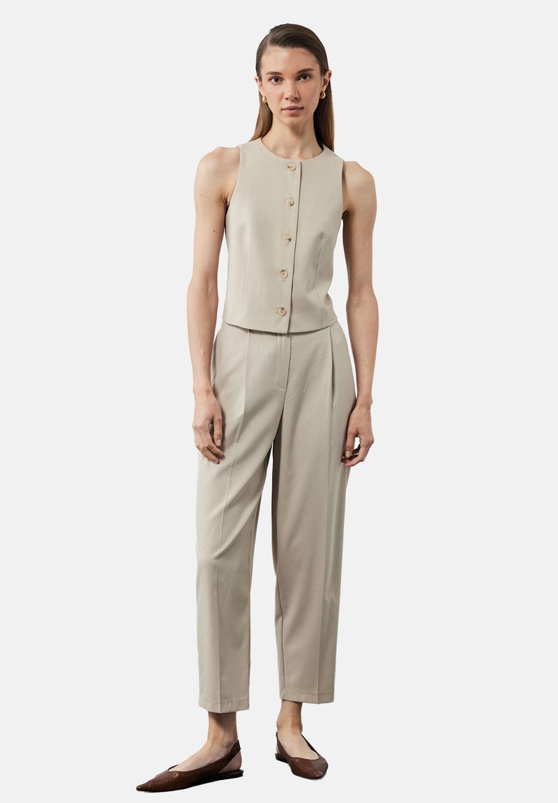 Top sin mangas color beige con botones en la parte delantera, combinado con pantalones de tiro alto y corte recto en un tono a juego; la modelo lleva zapatos planos marrones.